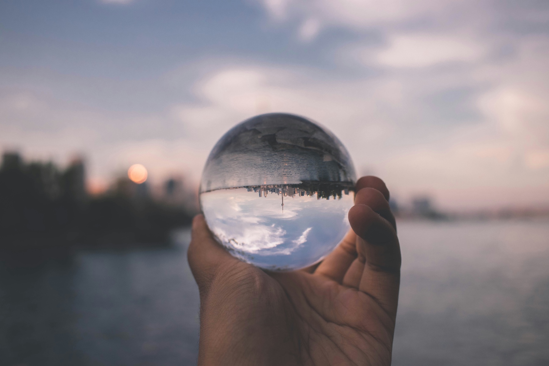 A hand holding a glass sphere that refracts a waterfront city at sunset